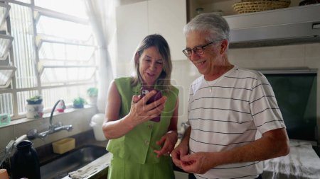Photo for Older couple looking at cellphone device together in kitchen. Wife showing screen to husband, authentic real life laugh and smile - Royalty Free Image