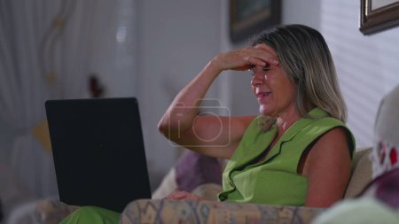 Foto de Mujer madura frustrada reaccionando negativamente a la notificación en frente de la computadora portátil sentada en el sofá de casa por la noche - Imagen libre de derechos