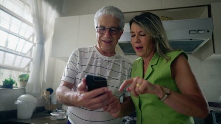 Téléchargez les photos : Couple âgé regardant appareil de téléphone portable ensemble tout en se tenant dans la cuisine. mari plus âgé montrant l'écran du téléphone à la femme d'âge moyen regardant le contenu en ligne - en image libre de droit