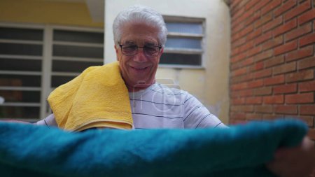 Téléchargez les photos : Homme âgé mettre des serviettes sur l'épaule après les avoir retirées des cordes à linge, faire des tâches ménagères domestiques - en image libre de droit