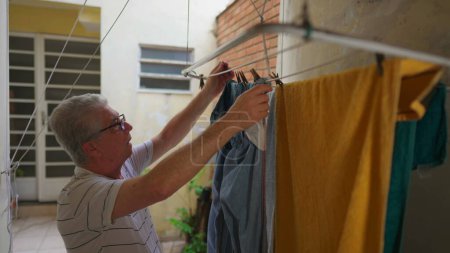 Téléchargez les photos : Homme âgé faisant des tâches ménagères domestiques, enlever les vêtements du cintre dans la cour arrière de la maison, résidence occasionnelle de la personne âgée - en image libre de droit