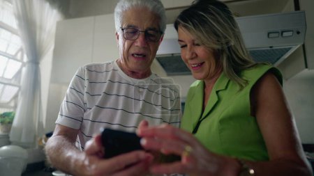 Téléchargez les photos : Couple âgé regardant appareil de téléphone portable ensemble tout en se tenant dans la cuisine. mari plus âgé montrant l'écran du téléphone à la femme d'âge moyen regardant le contenu en ligne - en image libre de droit