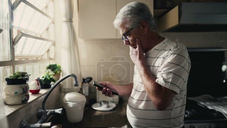 Foto de Hombre de edad avanzada pensativo mirando la pantalla del teléfono ponderando dilema y la toma de decisiones con un dispositivo de teléfono inteligente mientras está de pie en la cocina del hogar por la ventana - Imagen libre de derechos