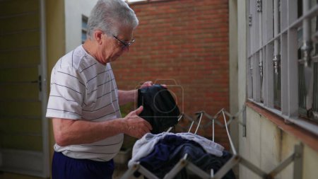 Photo for Senior man putting clothes to dry on hanger in home backyard. Independent elderly person doing domestic weekly routine at casual humble residence - Royalty Free Image