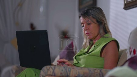 Téléchargez les photos : Femme âgée concentrée tapant sur ordinateur portable assis à la maison canapé dans la soirée. Femme caucasienne plus âgée engagée dans la technologie moderne, composant e-mail - en image libre de droit