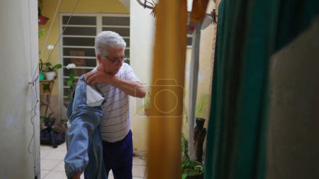 Téléchargez les photos : Senior homme retournant pantalon de l'intérieur après séchage sur cintre, personne âgée faisant la routine de corvée domestique en redressant les jeans - en image libre de droit