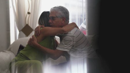 Téléchargez les photos : Homme âgé étreignant femme mûre dans un câlin empathique à la maison assis sur le canapé. Personnes âgées faisant preuve de soutien et d'aide, s'embrassant - en image libre de droit