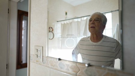 Photo for Senior man standing in front of bathroom mirror looking at his own reflection, contemplative elederly person starting the day morning routine - Royalty Free Image