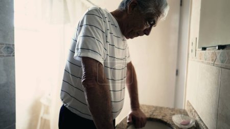 Photo for Depressed senior man standing by bathroom sink looking at his own reflection in mirror. Elderly person struggling with sorrow and regret - Royalty Free Image