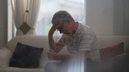 Téléchargez les photos : Déprimé Homme âgé souffrant seul à la maison aux prises avec des regrets et de l'anxiété. Candid personne âgée sentiment de frustration et de préoccupation assis à la maison sur le canapé - en image libre de droit