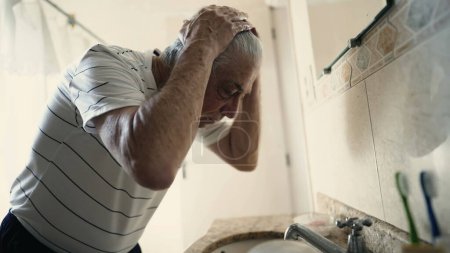 Téléchargez les photos : Homme âgé commençant la journée dans le rituel du matin debout à l'intérieur de la salle de bain en regardant son propre reflet devant le miroir - en image libre de droit