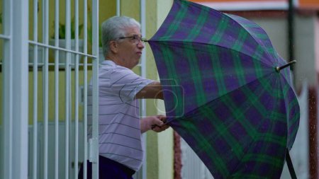Foto de Hombre mayor abriendo paraguas, sale a la calle bajo la lluvia, cierra la puerta principal detrás de él. Personas mayores que abandonan la residencia durante el día lluvioso en cámara lenta - Imagen libre de derechos