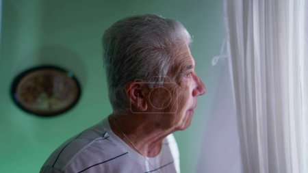 Senior man standing by window at home looking out view through curtains with pensive expression. Contemplative elderly gray-hair man watching neighborhood from residence