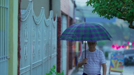 Téléchargez les photos : Homme âgé marchant sous la pluie tenant parapluie, personne âgée caucasienne se promenant dans le trottoir de la rue, marche en avant au ralenti, saison des pluies - en image libre de droit