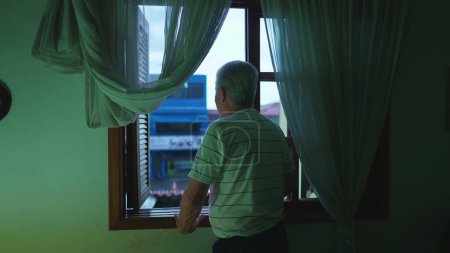 Foto de Hombre mayor de pie junto a la ventana del dormitorio por la noche viendo caer la lluvia de la tarde. Contemplativo anciano jubilado de pelo gris observando el vecindario desde casa - Imagen libre de derechos