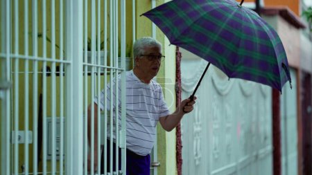 Téléchargez les photos : Un homme plus âgé sort sous la pluie, ouvre un parapluie et ferme la porte, marche sur le trottoir - en image libre de droit