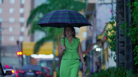 Téléchargez les photos : Femme pensive marchant sous la pluie tenant parapluie, se promenant dans un environnement urbain trottoir par mauvais temps. Personne marche vers l'avant dans la rue pendant l'après-midi pluvieux - en image libre de droit