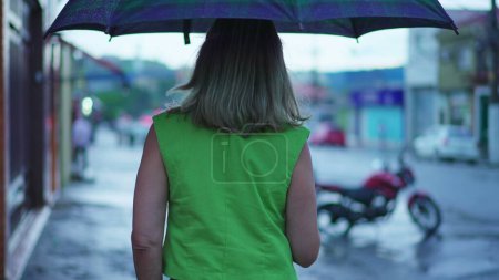 Foto de Detrás de la persona caminando bajo la lluvia sosteniendo el paraguas. Mujer paseando por la calle urbana durante la tarde de mal humor, lloviendo en cámara lenta - Imagen libre de derechos