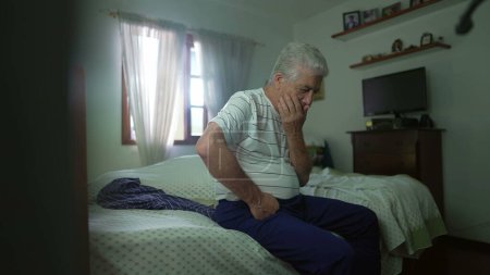 Téléchargez les photos : Homme âgé pensif assis au chevet du lit en contemplation. Retraité personne âgée réfléchissant à la décision avec la main dans le menton dans la réflexion mentale profonde dans la chambre décontractée - en image libre de droit