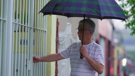 Photo for Elderly Individual Opens Gate and Enters Home, Guarded by Umbrella During Rain - Royalty Free Image