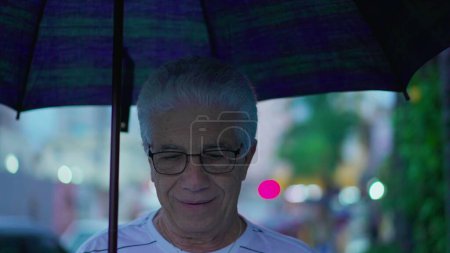 Foto de Un hombre mayor bajo la lluvia sosteniendo el paraguas. Anciano canoso maduro caucásico hombre camina hacia adelante en la acera de la calle de la ciudad, llega a casa - Imagen libre de derechos
