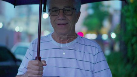 Téléchargez les photos : Un aîné sous la pluie tenant un parapluie. personne âgée cheveux gris mature caucasien mâle marche vers l'avant dans la rue de la ville trottoir, arrive à la maison - en image libre de droit
