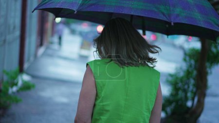 Téléchargez les photos : Retour de la personne marchant sous la pluie tenant parapluie. Femme se promenant dans la rue urbaine pendant l'après-midi lunatique, pleuvant au ralenti - en image libre de droit