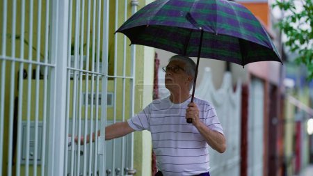 Téléchargez les photos : Un aîné rentre chez lui sous la pluie, déploie son parapluie et sécurise la porte, marche sur le trottoir - en image libre de droit