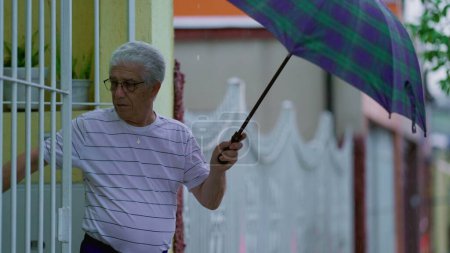 Foto de Hombre mayor abriendo paraguas, sale a la calle bajo la lluvia, cierra la puerta principal detrás de él. Personas mayores que abandonan la residencia durante el día lluvioso en cámara lenta - Imagen libre de derechos