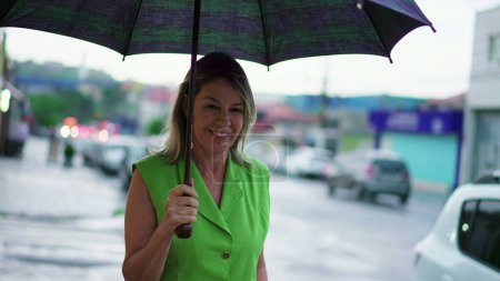 Téléchargez les photos : Heureuse femme d'âge moyen tenant un parapluie tout en marchant dans le trottoir de la rue de la ville de pluie - en image libre de droit