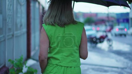 Téléchargez les photos : Retour de la personne marchant sous la pluie tenant parapluie. Femme se promenant dans la rue urbaine pendant l'après-midi lunatique, pleuvant au ralenti - en image libre de droit