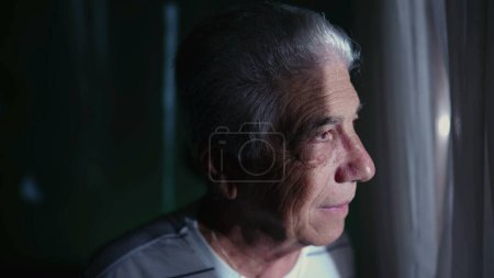 Téléchargez les photos : Homme âgé aux cheveux gris debout à la fenêtre de la maison, observant le voisinage avec expression contemplative - en image libre de droit