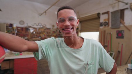 Foto de Un joven trabajador brasileño negro sonriente dentro del taller, parado por la máquina industrial y usando gafas protectoras, expresión gozosa en el trabajo - Imagen libre de derechos