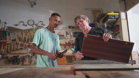 Foto de Maestro Carpintero trabajando en taller de carpintería con su joven aprendiz, mostrando cómo cortar madera con máquina. Joven apoyado con la ayuda de un mentor - Imagen libre de derechos
