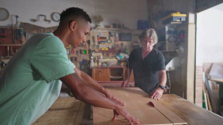 Téléchargez les photos : Scène d'atelier de menuiserie de maître charpentier et jeune apprenti travaillant à la menuiserie tranchant un morceau de bois avec une scie - en image libre de droit
