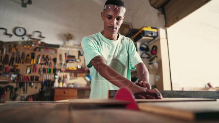 Foto de Joven carpintero que usa una sierra para cortar madera en el taller. Aprendiz enfocado en carpintería artesanal tienda - Imagen libre de derechos