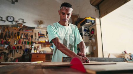 Photo for Concentrated Young Brazilian Carpenter Learning Profession in Workshop, using saw machine slicing wood - Royalty Free Image