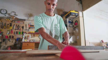 Foto de Un joven estudiante de carpintería negra midiendo madera, preparándose para usar una máquina de sierra con cinta métrica. Carpintero concentrado en el taller, profesión de aprendizaje - Imagen libre de derechos