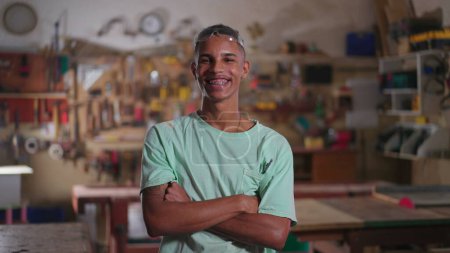 Foto de Retrato de un alegre carpintero brasileño sonriendo a la cámara con los brazos cruzados parado dentro del taller con herramientas y equipo colgando de fondo - Imagen libre de derechos