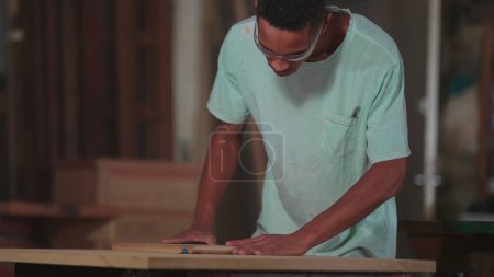 Foto de Un joven carpintero brasileño negro que trabaja en el taller de carpintería, recortando pieza de madera con máquina. Aprendiz concentrado comprometido con el trabajo - Imagen libre de derechos