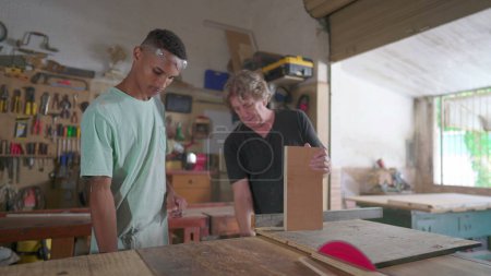 Téléchargez les photos : Processus d'enseignement dans l'atelier de menuiserie du maître charpentier et apprenti étudiant à l'aide d'une machine à scier - en image libre de droit
