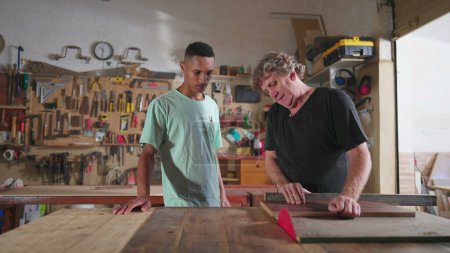 Photo for Carpenter and apprentice working at workshop, teacher orienting young man with saw machine at carpentry. Wood sawing scene of mentor teaching student - Royalty Free Image