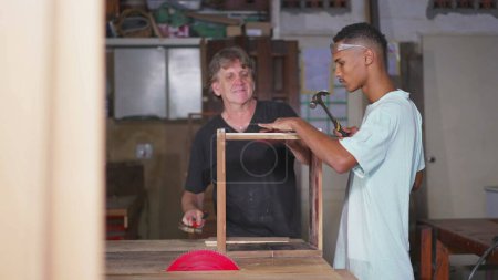 Foto de Master Carpenter Mentoring Aprendiz joven en la artesanía de muebles en el taller - Imagen libre de derechos