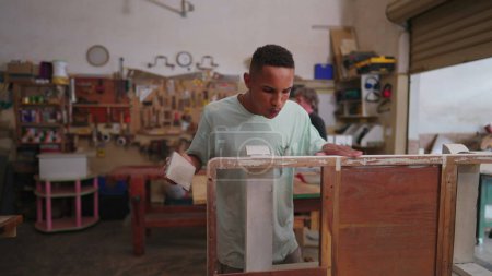Foto de Un joven carpintero negro puliendo muebles de madera de pie dentro del taller de carpintería. Trabajador concentrado reparando y creando objetos hechos a mano - Imagen libre de derechos