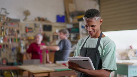Photo for Portrait of a young black Brazilian worker holding notepad and uniform inside carpentry workshop. Job occupation concept - Royalty Free Image