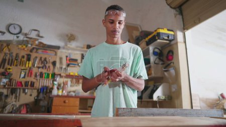 Foto de Un joven estudiante de carpintería negra midiendo madera, preparándose para usar una máquina de sierra con cinta métrica. Carpintero concentrado en el taller, profesión de aprendizaje - Imagen libre de derechos