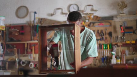 Foto de Participó en la escena artesanal de un joven carpintero brasileño negro que construye muebles en el taller, aprendiz con equipo de perforación, estudiante de carpintería de madera - Imagen libre de derechos