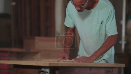 Photo for One young black Brazilian carpenter working at carpentry workshop, trimming piece of wood with machine. Concentrated apprentice engaged with work - Royalty Free Image