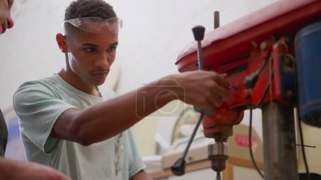 Téléchargez les photos : Maître charpentier enseignant à un jeune brésilien noir à utiliser une machine industrielle pour percer un trou dans l'écorce de bois à l'atelier de menuiserie - en image libre de droit