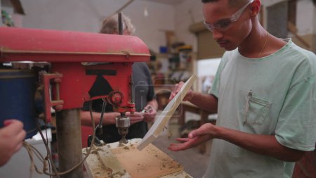 Foto de Maestro carpintero experimentado guiando a un joven aprendiz en el taller de carpintería. estudiante brasileño dando pulgares hasta jefe nexto a la máquina industrial - Imagen libre de derechos